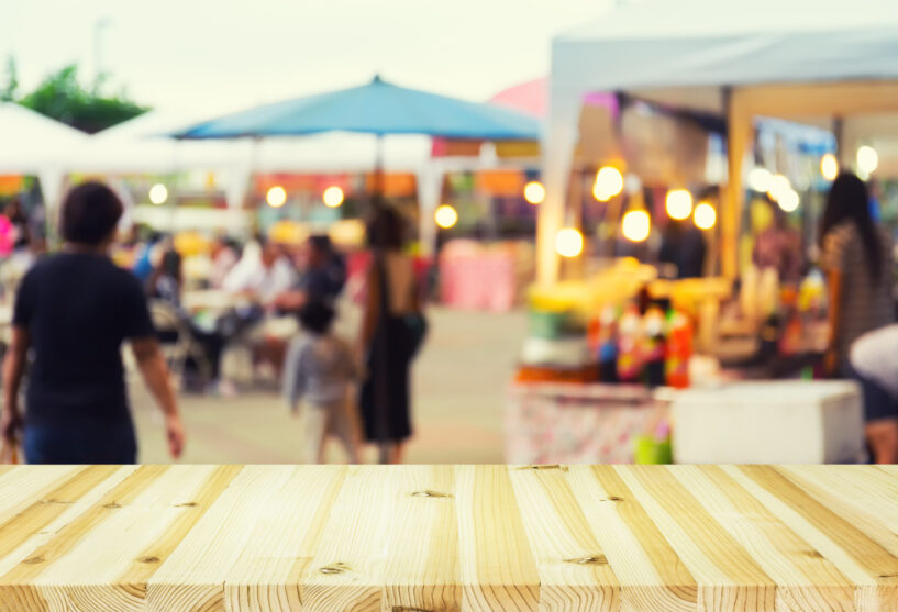 Blurred image of food fairs and food festivals consist of many booth and vendors at food stalls. People walking in street. Event in Chiang Mai at twilight montage with wood table top for background.