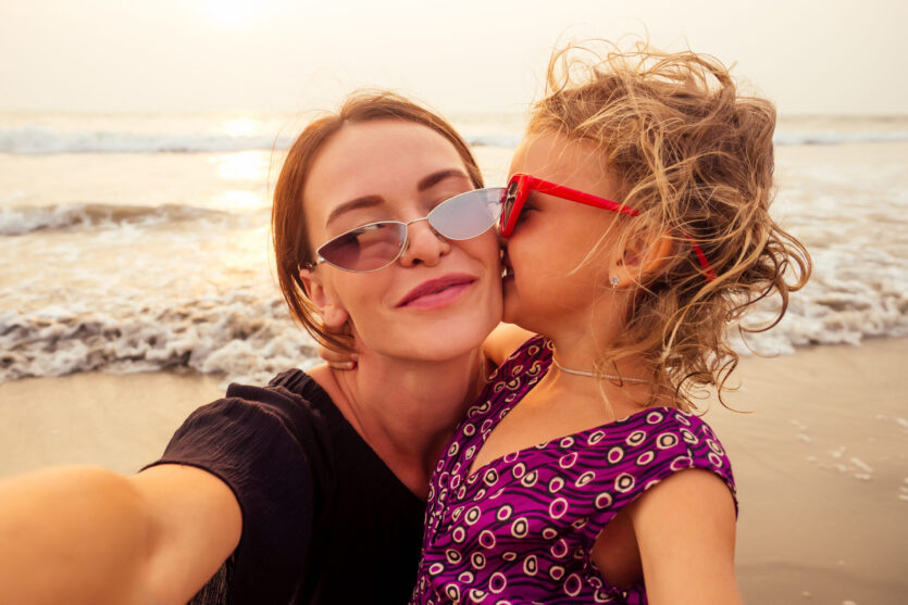Happy stylishly mother and daughter taking selfie at sandy beach on a sunset.mothers Day.little girl blonde and beautiful woman taking pictures on the phone tourism abroad online video calling